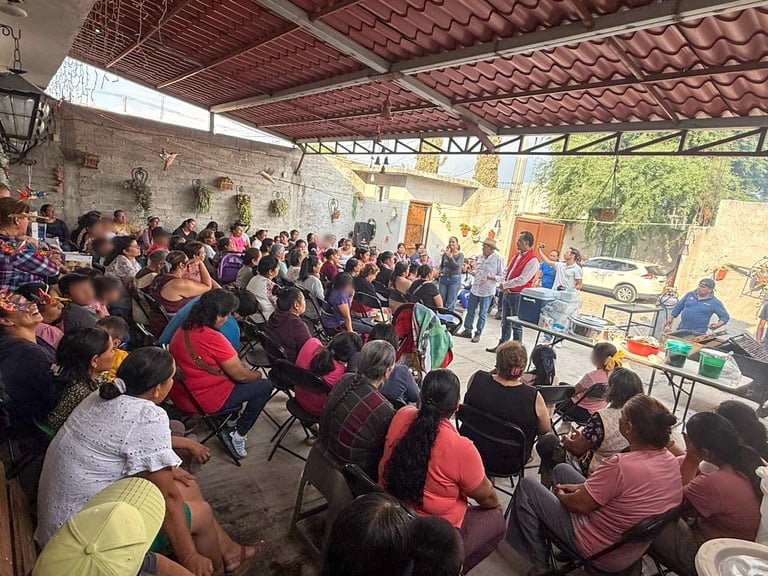 Large group of people seated and standing in a covered outdoor shelter or pavilion during what appears to be a community gathering or event