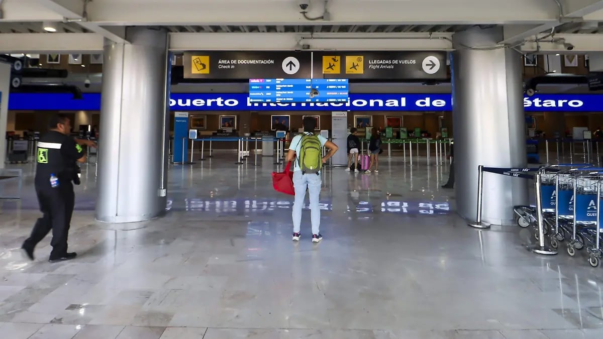 Person in yellow vest standing in empty airport terminal with blue signage, luggage carts visible on right side