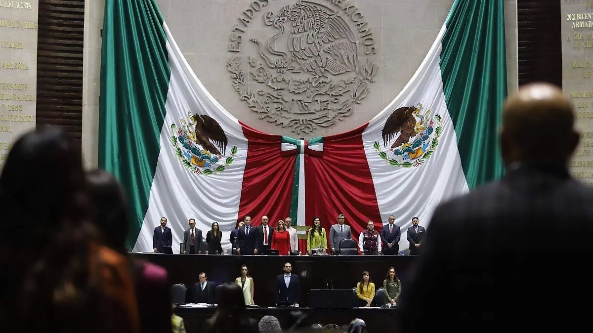 Mexican government officials on stage with the national seal and Mexican flag featuring green, white, and red colors during an official ceremony