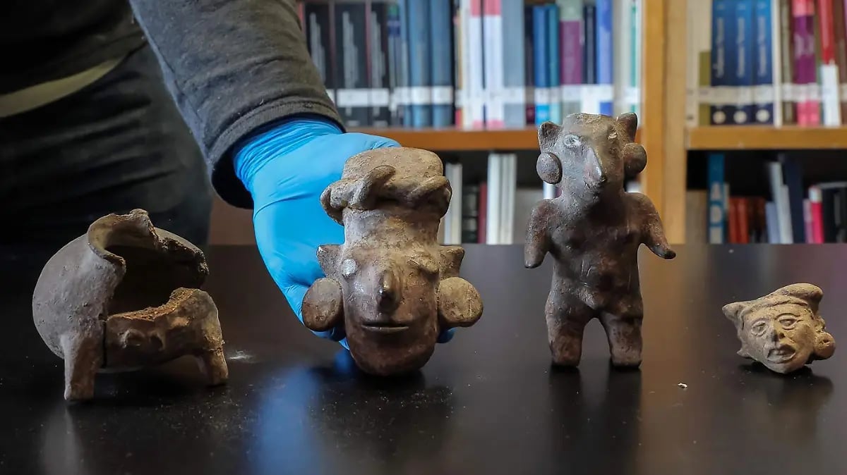 Hand in blue glove holding ancient clay figurines displayed on table with bookshelf in background