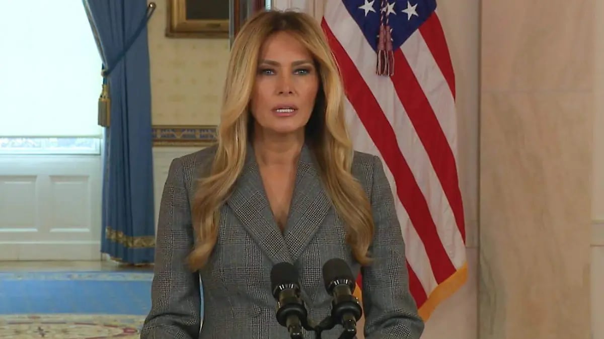 Woman in gray blazer speaking at podium with microphones in front of American flag in formal interior setting