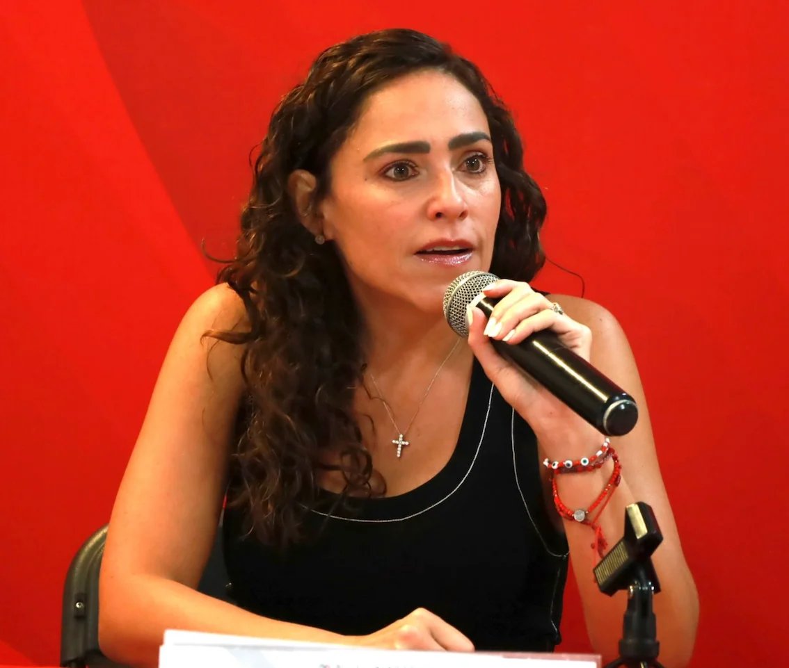 Woman with long dark wavy hair speaking into a microphone against a red background, wearing a black sleeveless top and cross necklace
