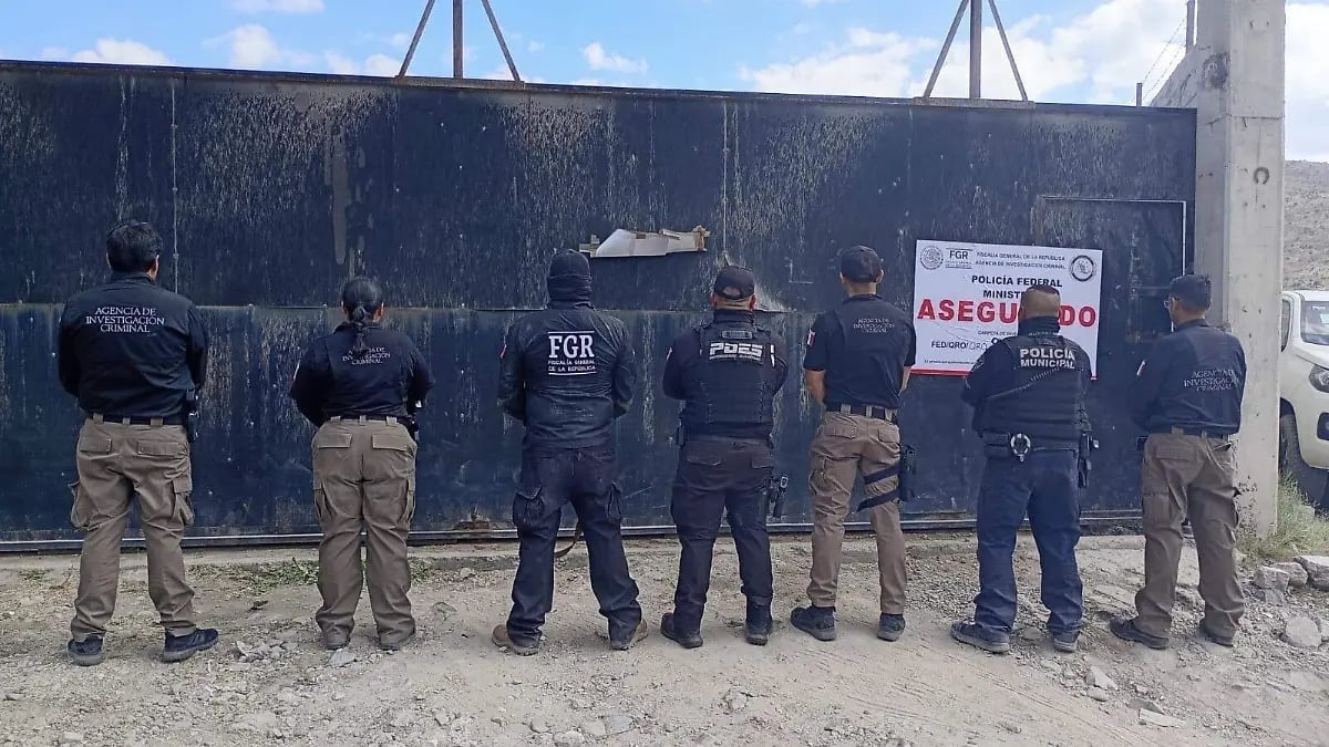 Seven law enforcement officers standing in a line facing a dark blue metal wall with signage, outdoors