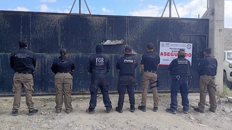Seven law enforcement officers standing in a line facing a dark blue metal wall with signage, outdoors