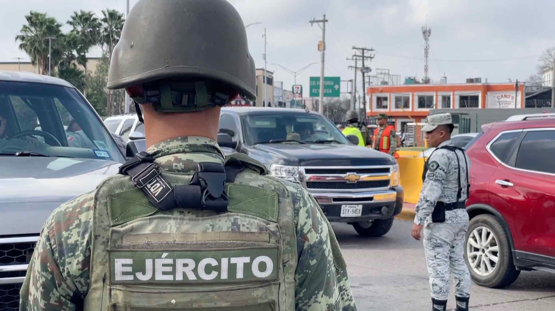 Mexican Army soldier from behind wearing camouflage uniform and helmet at a checkpoint with parked vehicles and personnel nearby