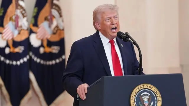 Older man with white hair wearing dark suit and red tie speaking at presidential podium with eagle seal