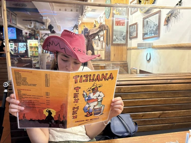 Person holding a Tijuana menu in a restaurant, wearing a pink cowboy hat, with warm interior decor in background