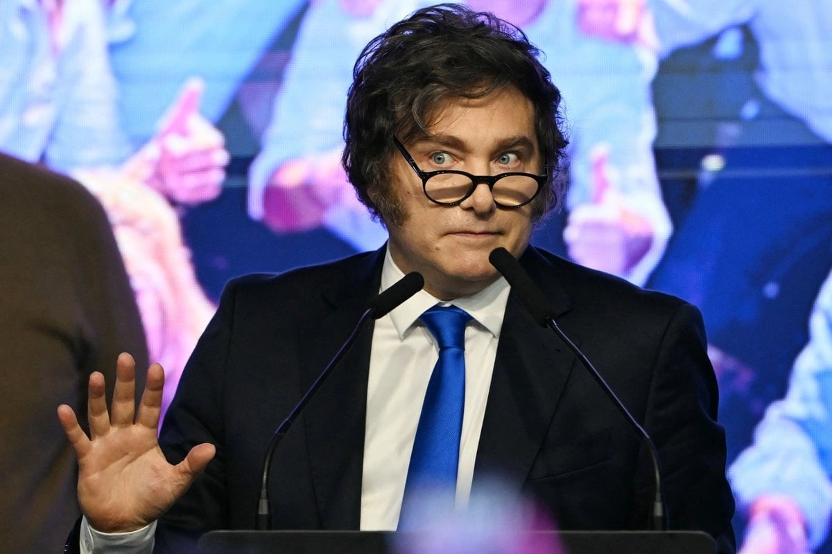 Man in black blazer and blue tie wearing glasses speaks at podium with colorful digital background